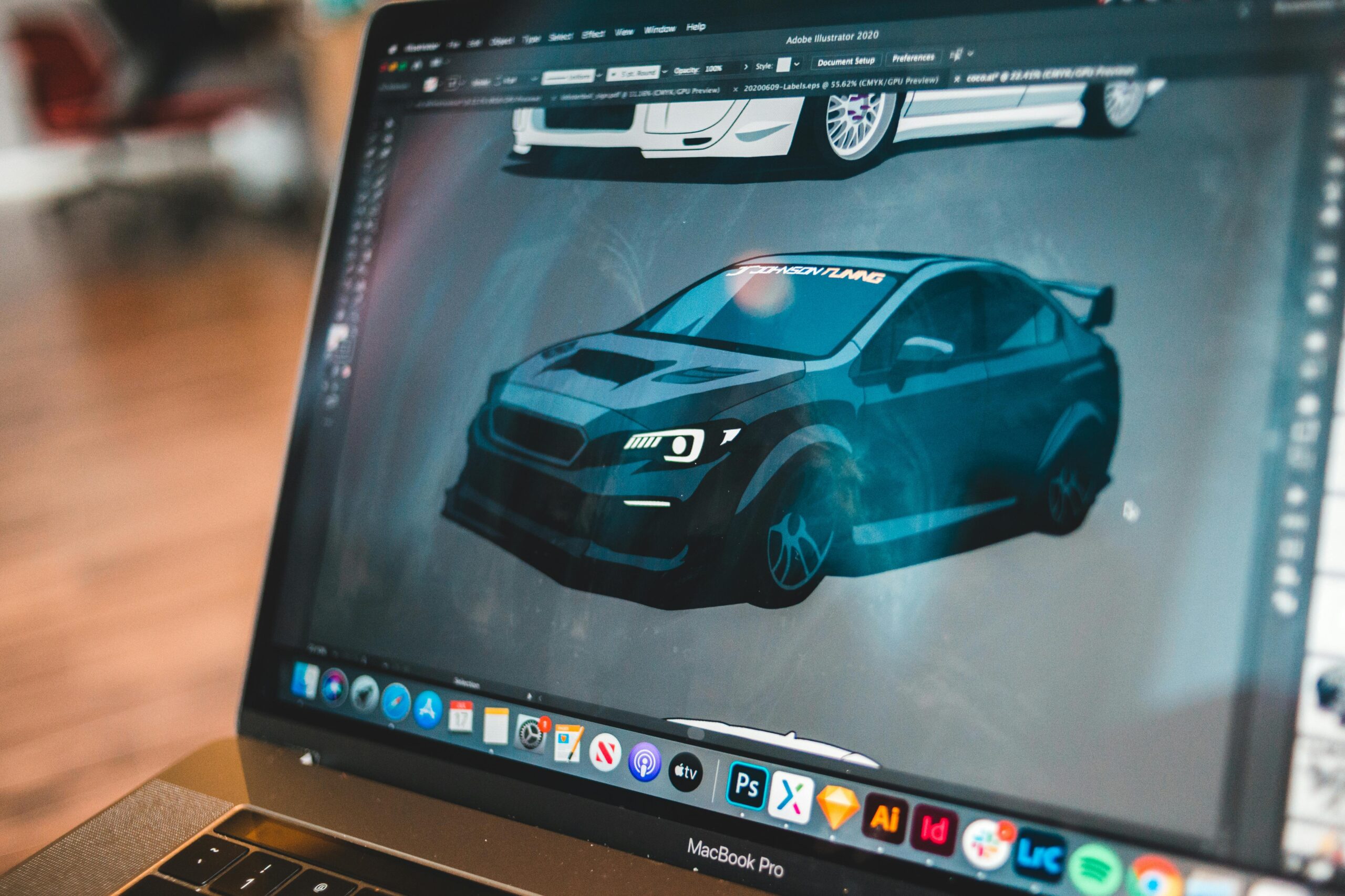 Un écran de MacBook Pro affichant Adobe Illustrator 2020. Le logiciel montre une illustration vectorielle détaillée d'une voiture de sport modifiée de couleur bleu foncé, avec une voiture blanche légèrement visible en arrière-plan. La barre des tâches macOS est visible en bas de l'écran.